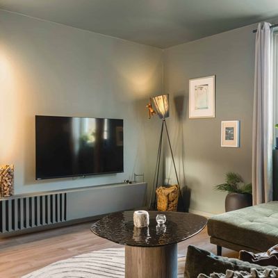 Stue med veggmontert TV, gulvlampe, grønn sofa og vindu mot skogen